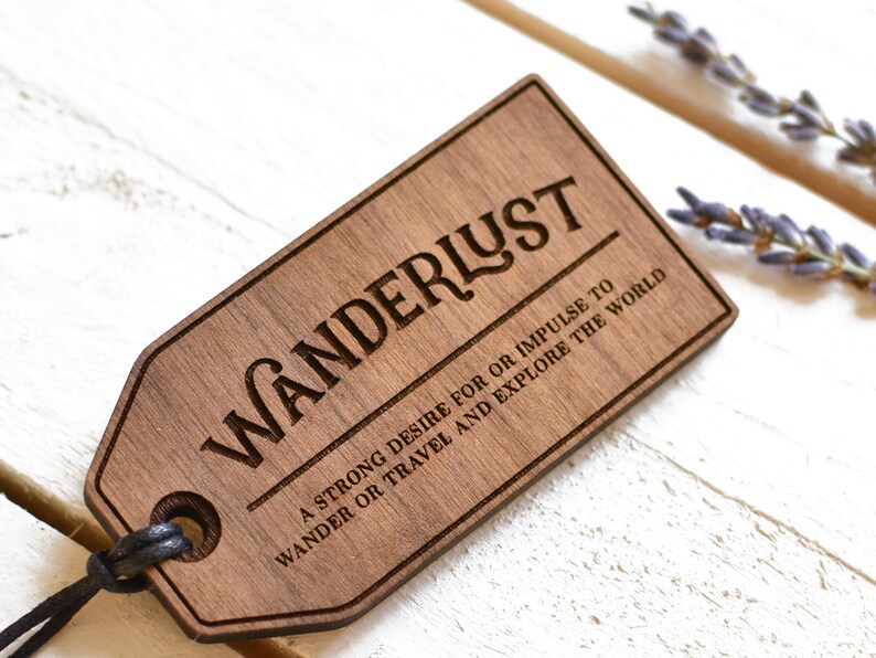 May include: A wooden tag with the word "WANDERLUST" engraved in a decorative font. The tag also includes the text "A strong desire for or impulse to wander or travel and explore the world."