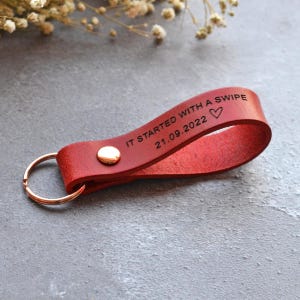 May include: A red leather keychain loop featuring a metallic rivet and a circular ring. The leather is engraved with the text IT STARTED WITH A SWIPE, the date 21.09.2022, and a heart symbol. The design is simple, functional, and durable.