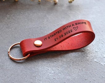 Puede incluir: Un llavero de cuero rojo en forma de bucle con un remache metálico y una anilla circular. El cuero está grabado con el texto IT STARTED WITH A SWIPE, la fecha 21.09.2022 y un símbolo de corazón. El diseño es sencillo, funcional y resistente.