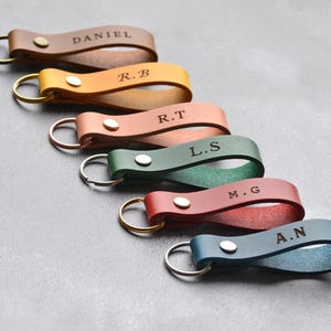 May include: A selection of personalised leather keyrings in shades of brown, yellow, pink, green, red, and blue. Each keyring has a silver or gold-coloured ring and is engraved with initials or a name. The keyrings are secured with a metal stud.