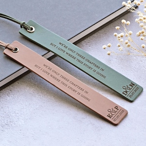 May include: Two leather bookmarks, one in a dusty rose hue and the other sage green, inscribed with the quote "WE'RE ONLY THREE CHAPTERS IN BUT I LOVE WHERE THIS STORY IS GOING". Each bookmark has a cord and eyelet. One is marked with "K&P" and "07.09.2018", the other with "D&N" and "25.07.2021".