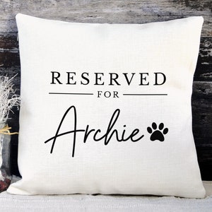 May include: White pillow with black text that reads "RESERVED FOR Archie" with a black paw print.