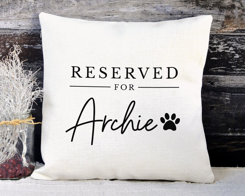 May include: White pillow with black text that reads "RESERVED FOR Archie" with a black paw print.