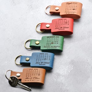 May include: Five leather keychains in tan, red, green, blue, and light brown, each featuring a metal ring, a snap button, and a laser-engraved calendar with a heart marking a specific date, along with names and text like "We said I Do" or "Engaged."
