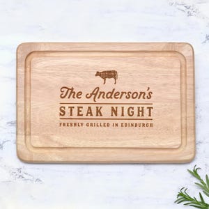 K&ouml;nnte beinhalten: Rechteckiges Holzschneidebrett mit geschnitztem Rand. Der Text "The Anderson's STEAK NIGHT" ist in einer dekorativen Schriftart eingraviert, zusammen mit einem Diagramm einer Kuh und dem Satz "FRESHLY GRILLED IN EDINBURGH".