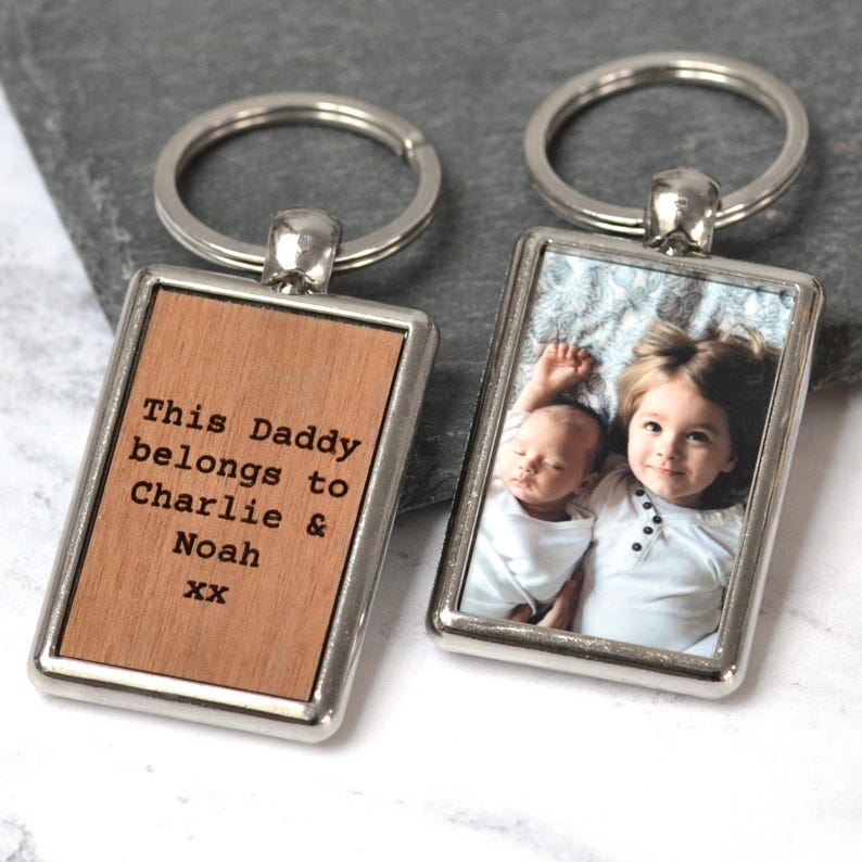 Puede incluir: Dos llaveros rectangulares de metal con marcos y anillas plateadas. Uno tiene una incrustaci&oacute;n de madera clara grabada con el texto This Daddy belongs to Charlie & Noah xx. El otro muestra una fotograf&iacute;a en color de dos ni&ntilde;os.