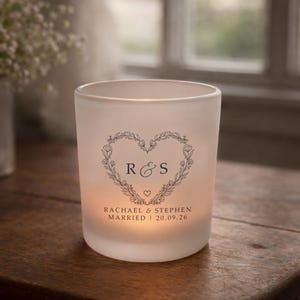 May include: A frosted glass candle holder with a floral heart design. The heart contains the initials "R & S" and a small heart. Below the heart, the text reads "RACHAEL & STEPHEN MARRIED 20.09.26".