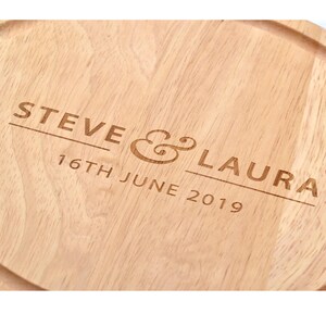 K&ouml;nnte beinhalten: Ein helles Holzschneidebrett mit eingraviertem Text. Der Text lautet "STEVE & LAURA" &uuml;ber "16TH JUNE 2019". Das Brett hat eine abgerundete Form und eine Nut am Rand.