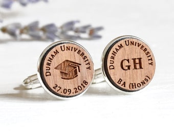 May include: A pair of wooden cufflinks with silver accents. One cufflink features the text "Durham University" with a graduation cap and the date "27.09.2018". The other cufflink features the text "Durham University" with the initials "GH" and the text "BA (Hons)".