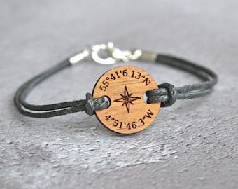 May include: A gray cord bracelet with a wooden charm featuring a compass and the coordinates "55°41'6.13"N 4°51'46.3"W".