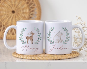 Könnte beinhalten: Zwei weiße Keramikbecher mit Illustrationen eines Hirsches und eines Bären, jeweils umgeben von einem grünen Kranz mit roten Beeren. Auf einem Becher steht "Mummy", auf dem anderen "Jackson".