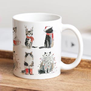 Könnte beinhalten: Weißer Keramikbecher mit Aquarellillustrationen von Katzen in festlicher Kleidung. Die Katzen tragen festliche Accessoires wie Schals, Geweihe und Lichterketten. Der Becher hat einen gebogenen Henkel und steht auf einer Holzoberfläche.