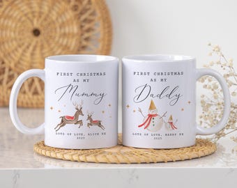 May include: Two white ceramic mugs with the text "First Christmas as my Mummy" and "First Christmas as my Daddy". The Mummy mug features deer, and the Daddy mug features snowmen. Both mugs have the year 2025.