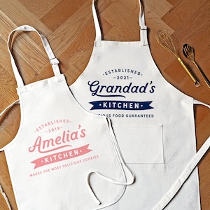 May include: Two white cotton aprons with black text. The top apron has the text "ESTABLISHED. -2021- Grandad's •KITCHEN.  OUS FOOD GUARANTEED". The bottom apron has the text "ESTABLISHED. -2019- Amelia's •KITCHEN. MAKES THE MOST DELICIOUS COOKIES".