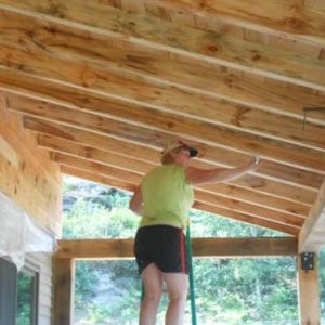 May include: A person wearing a green shirt and black shorts is painting the wooden beams of a porch. The porch has a wooden railing and a white plastic tarp hanging from the ceiling.