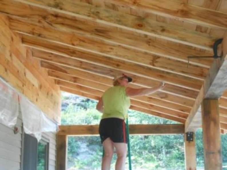 May include: A person wearing a green shirt and black shorts is painting the wooden beams of a porch. The porch has a wooden railing and a white plastic tarp hanging from the ceiling.