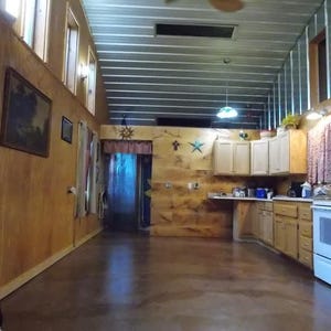 May include: A kitchen with wood paneling on the walls and a wood floor. The kitchen has white cabinets, a white stove, and a stainless steel sink. There are two windows on the left side of the room and a door leading to another room.