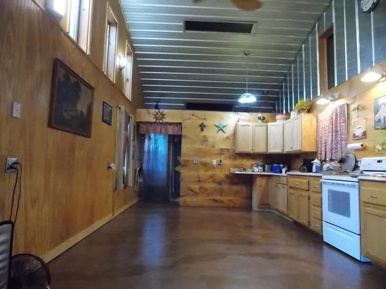 May include: A kitchen with wood paneling on the walls and a wood floor. The kitchen has white cabinets, a white stove, and a stainless steel sink. There are two windows on the left side of the room and a door leading to another room.
