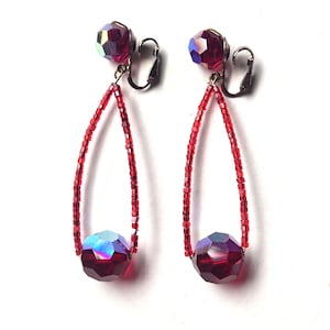 May include: A pair of clip-on earrings featuring a teardrop-shaped loop constructed from small red seed beads. Each earring is accented with two faceted, iridescent dark red crystal beads, one at the top near the clip and one at the base.