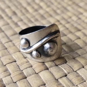 May include: A wide silver-toned metal ring featuring a smooth, polished surface with a diagonal raised band. The design includes three spherical protrusions of varying sizes integrated into the band, creating a textured, dimensional appearance.