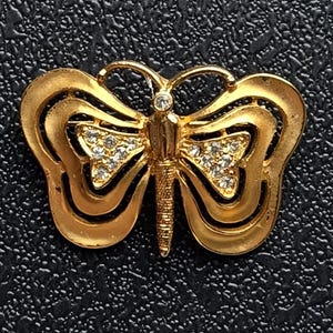 May include: A gold-toned butterfly brooch featuring openwork wings with smooth, curved edges. The center of the butterfly is textured, and the upper wing sections are inlaid with small, clear, sparkling stones. A single clear stone sits at the top.