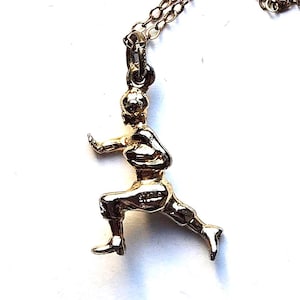 May include: A gold-colored pendant necklace featuring a small, detailed figure in a dynamic pose, possibly engaged in a martial art or exercise. The figure is attached to a delicate gold chain, creating a simple yet elegant accessory.