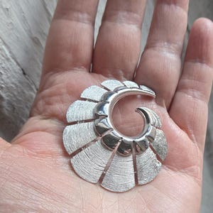Puede incluir: Broche plateado con diseño en forma de abanico. La broche presenta un elemento central curvado y segmentos texturizados que irradian. Se sostiene en una mano, mostrando su tamaño y detalle. La broche tiene un acabado metálico pulido.