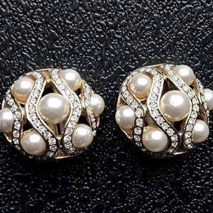 Puede incluir: Un par de pendientes redondos dorados con clip. Cada pendiente presenta una perla central rodeada de perlas más pequeñas y brillantes pedrería transparente. Los pendientes tienen un diseño vintage ornamentado.