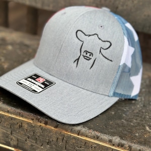 Può includere: Un cappello da camionista grigio e bianco con un design a bandiera americana rosso, bianco e blu sul retro. La parte anteriore del cappello presenta una silhouette di mucca ricamata in nero.