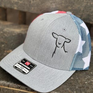 Può includere: Un cappello da camionista grigio e blu con un motivo a stelle bianche e una piccola pecora ricamata sul davanti. Il cappello ha un cinturino regolabile nero con un'etichetta che recita "Richardson 112 Trucker Adjustable".