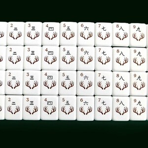 Peut inclure: Un ensemble de tuiles de Mahjong blanches avec des symboles et des chiffres bruns. Les tuiles pr&eacute;sentent des chiffres chinois et des motifs de bois de cerf. Le mot "CRAK" est imprim&eacute; sur certaines tuiles. Elles sont dispos&eacute;es sur un fond vert fonc&eacute;.