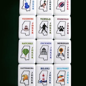 Peut inclure: Ensemble de tuiles blanches avec des illustrations et des noms de villes du Mississippi, dont Clarksdale, Greenwood, Oxford, Vicksburg, Tupelo, Starkville, Natchez, Jackson, Meridian, Hattiesburg, Biloxi et Gulfport. Chaque tuile porte le mot "JOKER" en rouge.