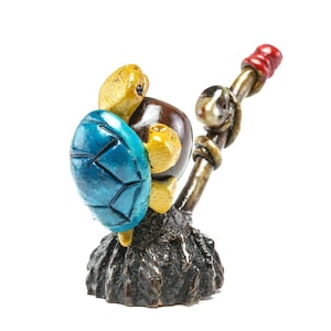 May include: A whimsical figurine of a turtle with a blue shell, brown body, and yellow head and limbs. The turtle holds a staff with a red tip. The base is textured and dark brown.