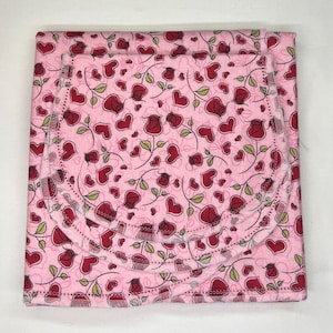 May include: A folded pink fabric with a repeating pattern of red roses, hearts, and green leaves. The fabric has a scalloped edge and is likely a baby blanket or similar item. The overall design is romantic and floral.