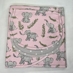 May include: Pink baby blanket with a gray elephant pattern and green leaf accents. The blanket has a gray border with white polka dots. The blanket is folded and ready to use.