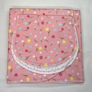 May include: A pink baby blanket and bib set. The set features a floral pattern with red, yellow, blue, and white flowers. The bib has a curved shape with a white border. The blanket is folded and the bib is placed on top.