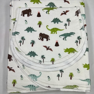 May include: A white baby blanket with a dinosaur and palm tree print. The blanket has a white border and is folded in thirds.