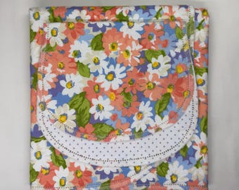 May include: A folded fabric item featuring a vibrant floral pattern with coral, white, and blue flowers on a light blue background. The piece includes a curved, white, polka-dot fabric accent along the bottom edge, finished with stitched edges.