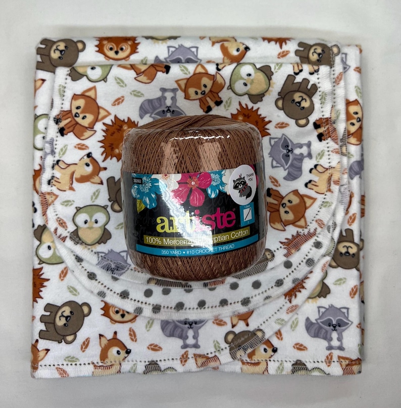 Puede incluir: Una bobina marr&oacute;n de hilo de ganchillo Artiste 100% algod&oacute;n egipcio mercerizado se encuentra sobre una manta blanca para beb&eacute; con un estampado de animales del bosque. La manta presenta im&aacute;genes de zorros, osos, mapaches y b&uacute;hos.
