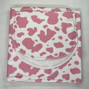 May include: A folded white blanket with a pink cow print. The blanket has a rounded edge with a stitched border. The pink spots are irregular in shape and size. The blanket is made of a soft material.