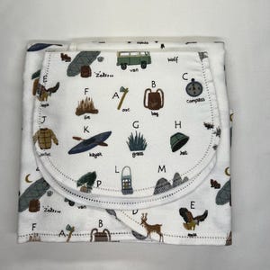 May include: White baby blanket with a colorful alphabet print featuring camping and outdoor themed illustrations. The blanket has a pocket on the front with a white trim.