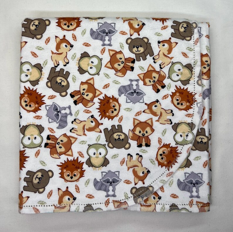 Puede incluir: Una manta para beb&eacute; blanca con un estampado repetido de animales del bosque, incluyendo osos, zorros, mapaches, b&uacute;hos y erizos. La manta tiene un borde doblado y est&aacute; decorada con peque&ntilde;os acentos de hojas.