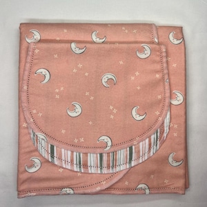 May include: A folded, coral-colored baby blanket and burp cloth set. The fabric features a repeating pattern of smiling crescent moons and small starbursts. The burp cloth has a striped border in shades of green, gray, and white.