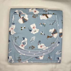 May include: A baby blanket and burp cloth set. The set features a light blue background with a repeating pattern of white teddy bears, flowers, and foliage. The burp cloth is folded over the blanket, with a rounded edge and a light blue trim.