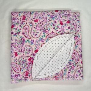 May include: A folded baby blanket with a pink paisley pattern. The blanket features a white, leaf-shaped accent with gray polka dots. The paisley design includes shades of pink, purple, and green.
