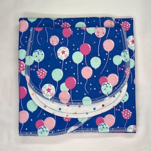 May include: A square, folded baby blanket with a blue background and a pattern of pink, mint green, and white balloons. The blanket has a white trim with a dotted pattern and a stitched edge.