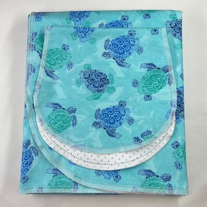 May include: A blue and green baby blanket with a sea turtle pattern. The blanket is folded in thirds and has a white polka dot trim.