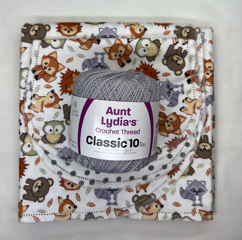 Puede incluir: Hilo de ganchillo Aunt Lydia's Classic 10 gris, sobre una manta para beb&eacute; con estampado de animales del bosque. La manta presenta varias criaturas del bosque en marr&oacute;n, gris y naranja sobre un fondo blanco.