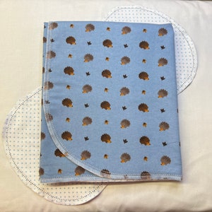 May include: A baby blanket and two burp cloths. The blanket is light blue with a repeating pattern of brown hedgehogs, acorns, and leaves. The burp cloths are white with blue polka dots and a scalloped edge.