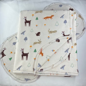 May include: A cream-colored baby blanket with a woodland animal print. The blanket features deer, foxes, squirrels, and birds. The blanket is folded in half and has a scalloped edge.